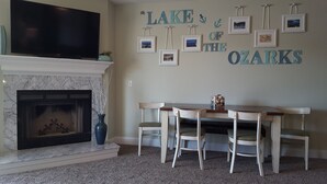 Una sala de estar con chimenea, un televisor montado encima, una mesa de comedor con sillas, y una pared con cuadros y el texto "LAKE OF THE OZARKS".