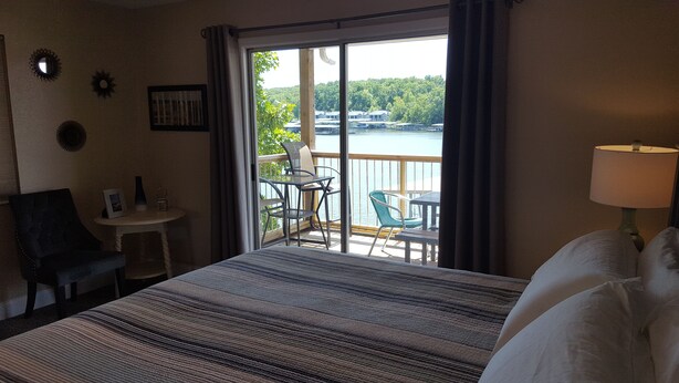 Habitación de hotel con cama, escritorio, silla y vista a un lago a través de un ventanal grande.