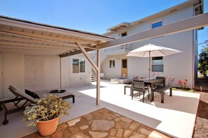 A covered outdoor patio with a stone floor, a table with chairs, a lounge chair, and a potted plant.