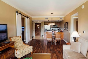 A living room with a TV, a sofa, a dining area with chairs, a kitchen with appliances, and a hallway leading to another room. - KLVR presents Honua Kai K612*Two bedrooms*one bath (Lahaina)