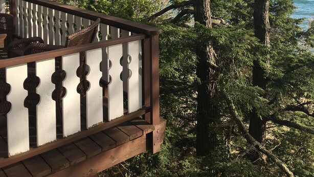 Une terrasse en bois, agrémentée de meubles en osier, donnant sur une zone côtière bordée d’arbres et offrant une vue sur la mer.