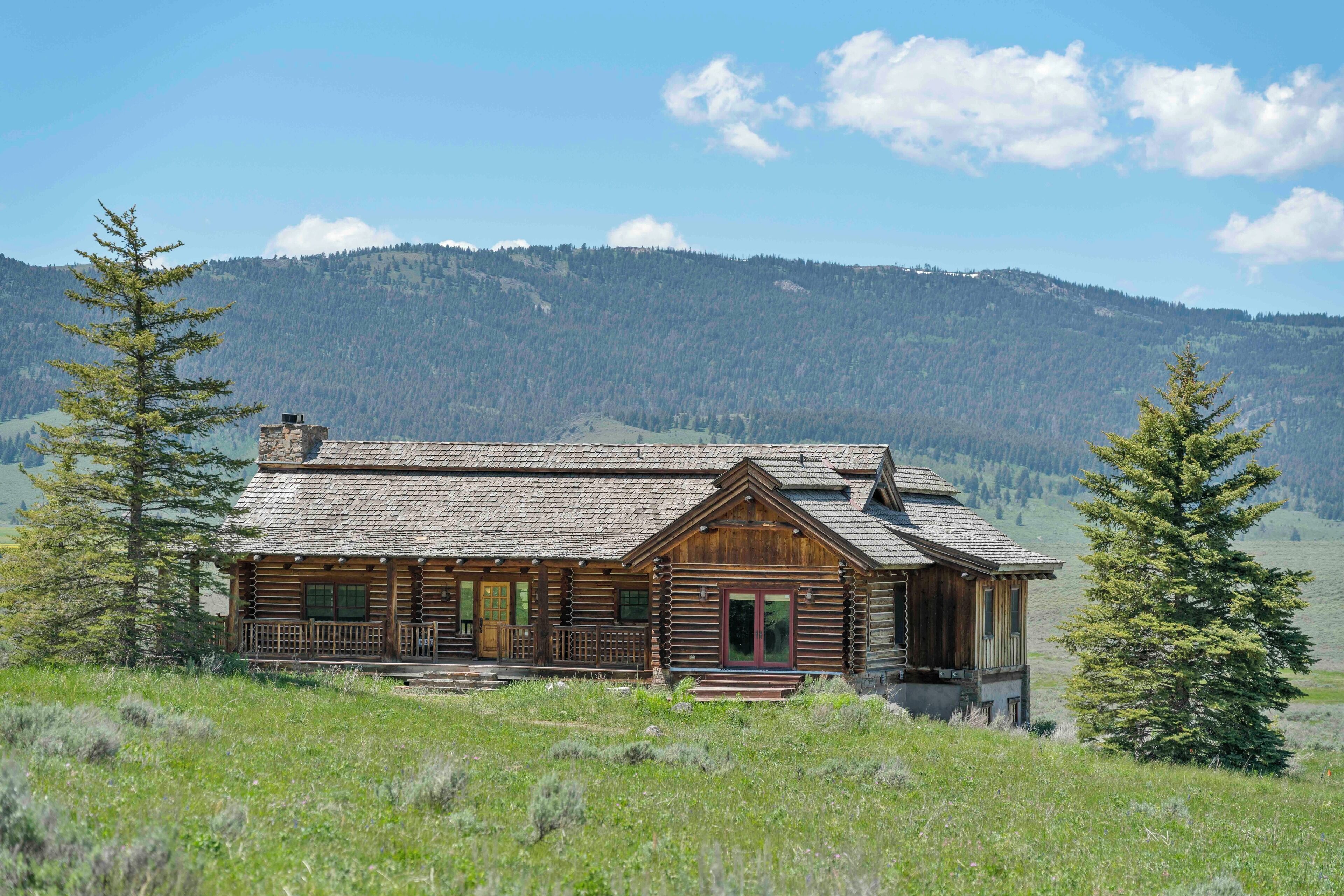 A log cabin with a porch, surrounded by a grassy field and a forested mountain backdrop.