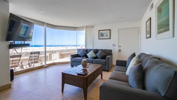 A living room with a large window, a flat-screen TV, a sofa, a coffee table, and a view of the beach. - 50' of Ocean Front * Exclusive Ground Level Patio * AC (San Diego)
