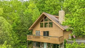 Eine Holzhütte mit einem Steinofen, einem Balkon und umgeben von dichtem Wald.