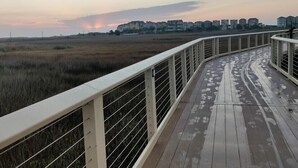 Una pasarela de madera con barandillas metálicas, que conduce a una zona con césped y edificios al fondo.