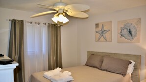 Una cama bien hecha con cabecera, ventilador de techo y dos cuadros.