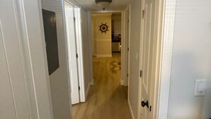 Un pasillo estrecho con paredes blancas y pisos de madera, que conduce a una habitación con una puerta visible y una rejilla en el techo.