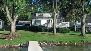 A residential property with a dock extending into a body of water, surrounded by trees and a well-maintained lawn. - Beautiful Lake Front Home On Como with boat and motor included (Lake Geneva)