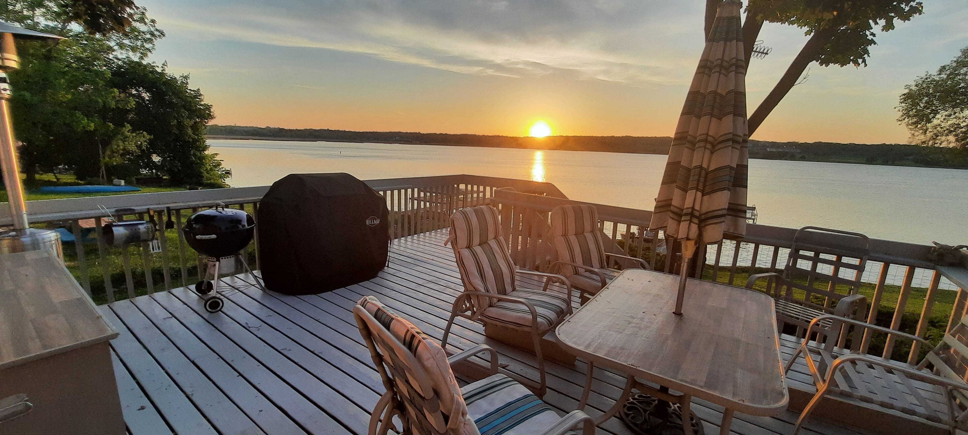 Une terrasse dotée de mobilier d’extérieur, d’un barbecue et offrant une vue sur le coucher de soleil au-dessus d’une étendue d’eau.