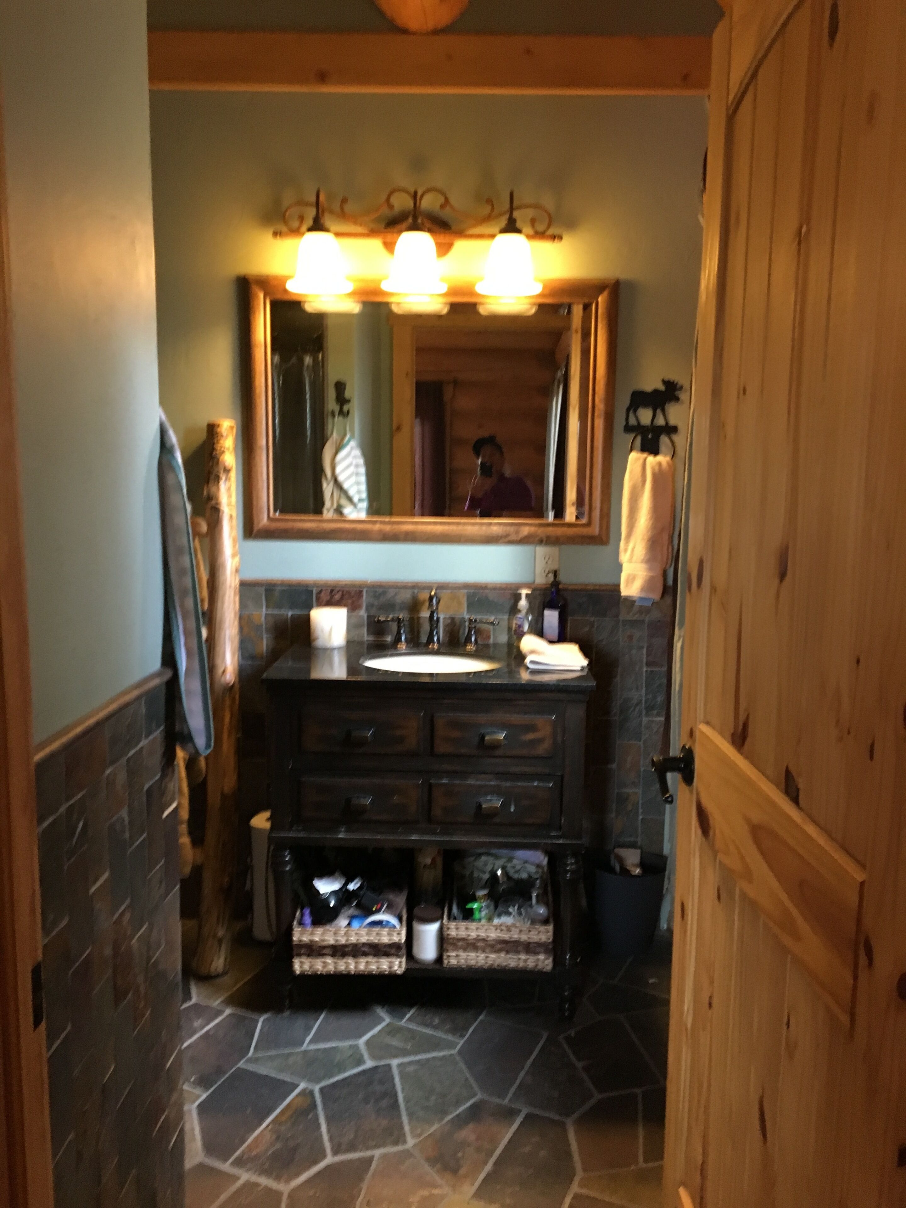 A bathroom with a stone floor, wooden vanity, and a large mirror with three lights.