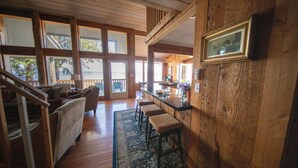 A spacious living area with a wooden staircase, a bar area with stools, and large windows offering a view of the outdoors.