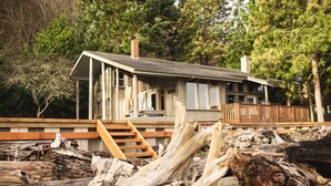 A wooden cabin with a deck, surrounded by trees and driftwood.