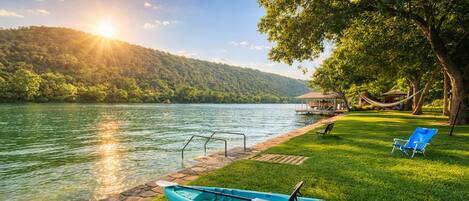 Un paisaje junto al lago con un kayak, un muelle y una zona verde con una silla.