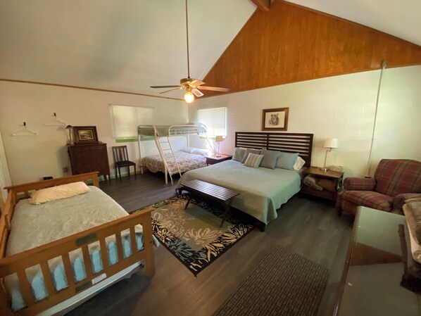 Un dormitorio con una cama doble, una cama individual, cabecera de madera, ventilador de techo, mesita de noche, una silla y una alfombra.