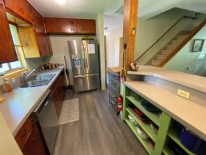 A kitchen with wooden cabinets, a stainless steel refrigerator, a sink, and a green shelf with dishes.