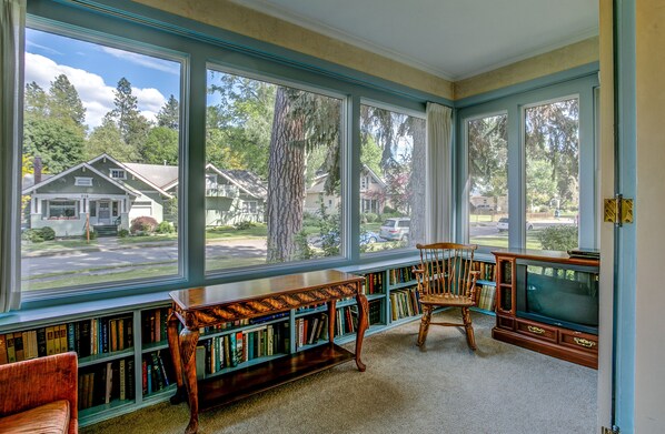 Une véranda avec de grandes fenêtres, une bibliothèque en bois, un téléviseur et un fauteuil.