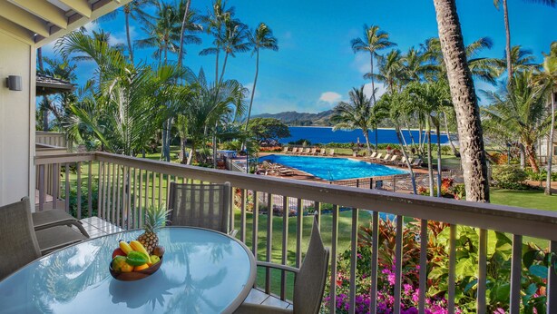 Un balcone con un tavolo in vetro, una ciotola di frutta e vista su una piscina circondata da palme e una catena montuosa.