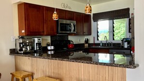 Una cucina moderna con mobili in legno, un piano di lavoro in granito, una macchina per il caffè, un bollitore e un forno integrato. Sono presenti tre sgabelli di legno e una finestra con vista sul verde.