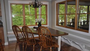 Comedor con mesa y sillas de madera, ventanas amplias con vista al bosque y una araña.