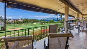 A balcony with a view of a golf course, outdoor seating, and a covered area with chairs and tables. - WOW- New 2bd/2ba, Largest Lanai w/Ocean & Golf Course Views, Lowest Resort Fee! (Lahaina)