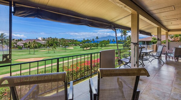 A balcony with a view of a golf course, outdoor seating, and a covered area with chairs and tables.