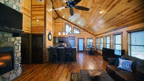 Un intérieur de cabane spacieux, doté d’une cheminée en pierre, d’un coin cuisine, d’un espace repas et d’un salon avec un canapé et un téléviseur.