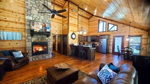 L’intérieur d’une cabane en rondins, avec une cheminée en pierre, des murs en bois, un ventilateur de plafond, un coin cuisine et un espace salon avec une table basse.
