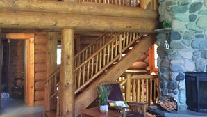 A spacious log cabin interior with a stone fireplace, wooden staircase, and a cozy seating area. - Loons Nest - Cozy, secluded 4 bedroom log cabin in the heart of the wilderness! (Tyaughton Lake)