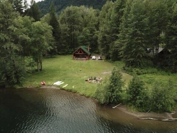 A cabin surrounded by dense forest, a small clearing, and a body of water. - Loons Nest - Cozy, secluded 4 bedroom log cabin in the heart of the wilderness! (Tyaughton Lake)