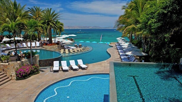 Un espace piscine agrémenté de chaises longues et de parasols, entouré de palmiers et offrant une vue sur l’océan.