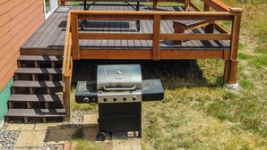 Una terraza de madera con una parrilla, mesas de picnic y una escalera que lleva a un edificio.