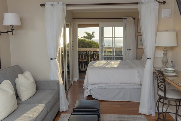 A bedroom with a bed, a grey sofa, a small table with a lamp, and a view of the outdoors.