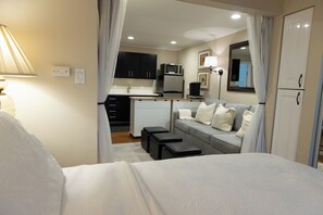 A compact hotel room with a bed, a small kitchenette, a sofa with pillows, and a mirror.