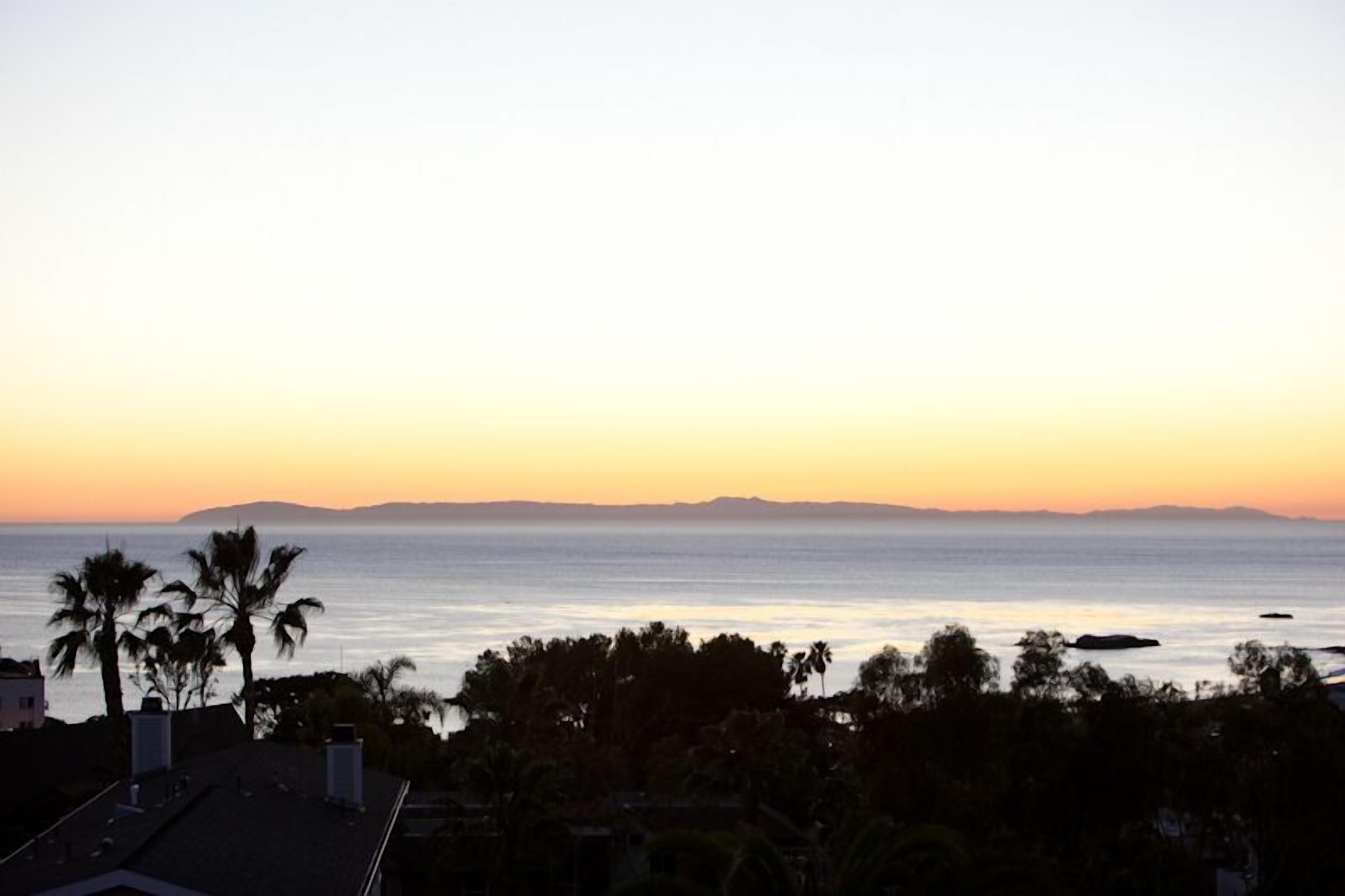A coastal view at sunset with palm trees and distant mountains.