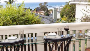 Un balcón con una mesa y sillas de vidrio, con vista a la costa, casas y palmeras.