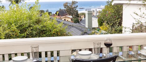 Un balcón con una mesa y sillas de vidrio, con vista a la costa, casas y palmeras.