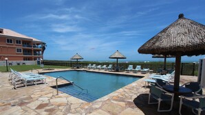 Ein Swimmingpool mit Liegestühlen und Reetdach-Sonnenschirmen, umgeben von Steinplatten und einem Gebäude im Hintergrund.