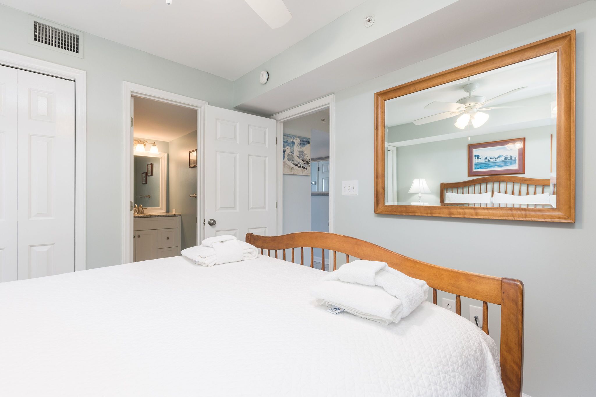 Une chambre à coucher avec un grand lit, une tête de lit en bois, un ventilateur de plafond, un miroir et une porte donnant sur une autre pièce.