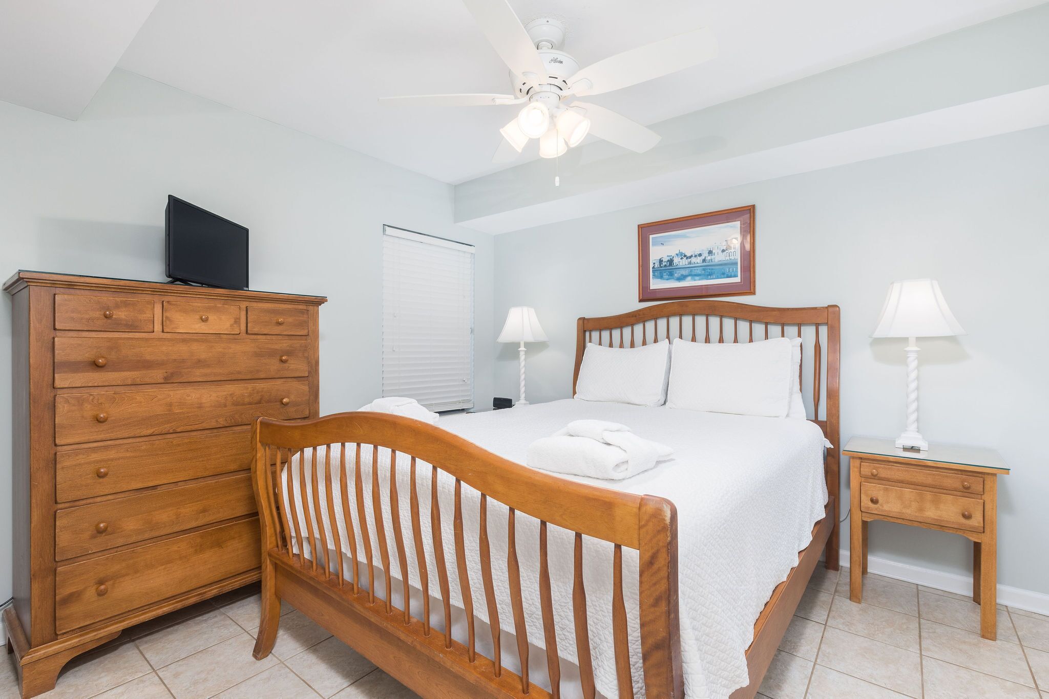 Une chambre à coucher avec un lit en bois, une table de chevet avec une lampe, une commode et un ventilateur de plafond.