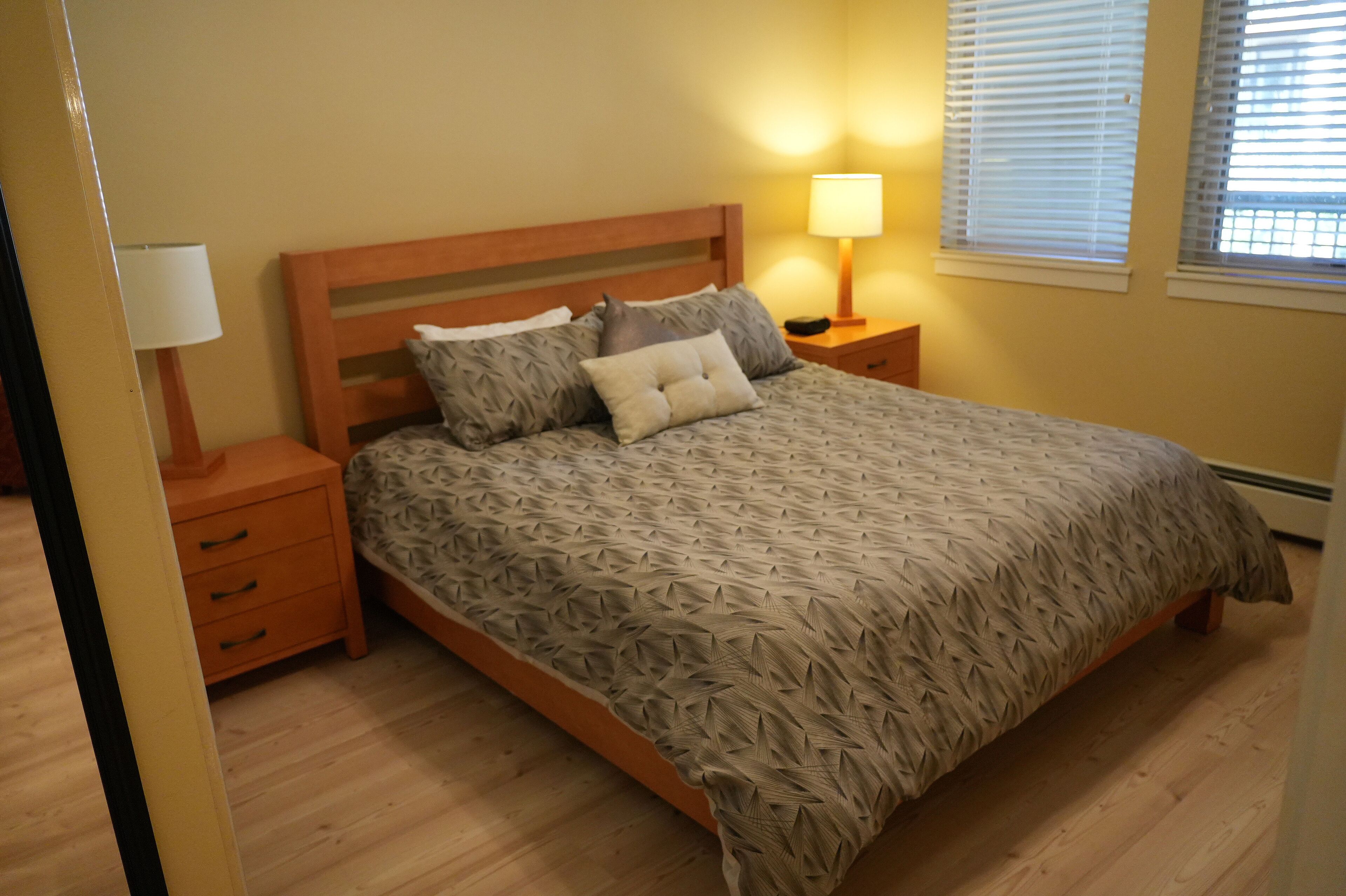 A bedroom with a wooden bed, a nightstand with a lamp, and a window with blinds.