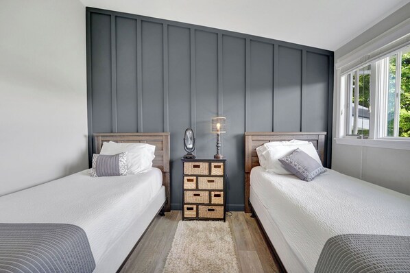A bedroom with two beds, a nightstand with a lamp, and a window with a view of greenery.