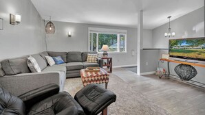 A modern living room with a grey sectional sofa, a black leather chair, a patterned ottoman, a television, and a large window providing natural light. - Beautiful and modern 3 bedroom, 1 bath Duplex.  Sleeps 6 (Chaffee)