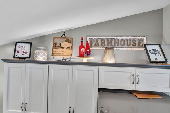 A kitchen with white cabinets, a farmhouse sign, and decorative items including a framed sign saying "Let's go Buffalo" and a bottle of wine.
