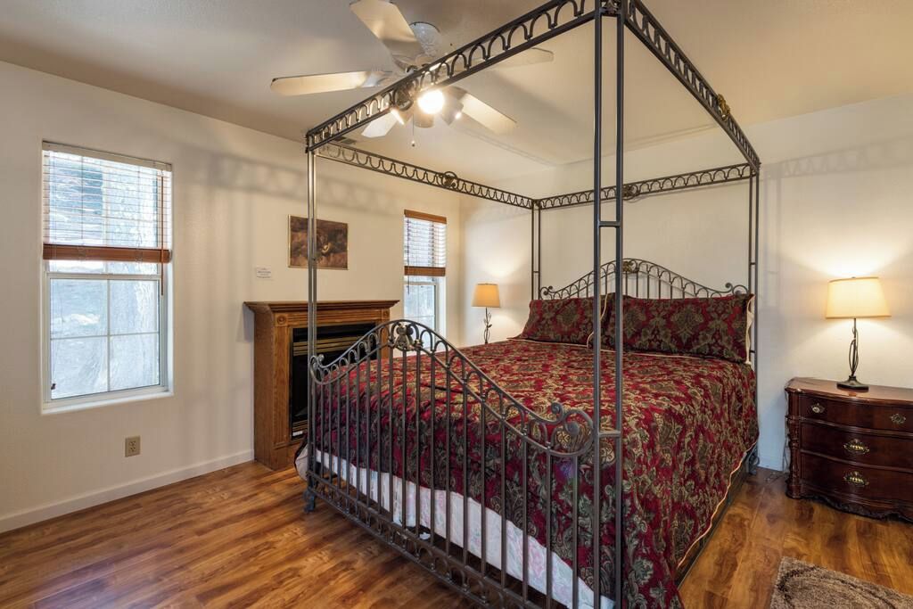 Een hemelbed met een rode en gouden spreideken, een houten nachtkastje met een lamp, en een bureau met een lamp.