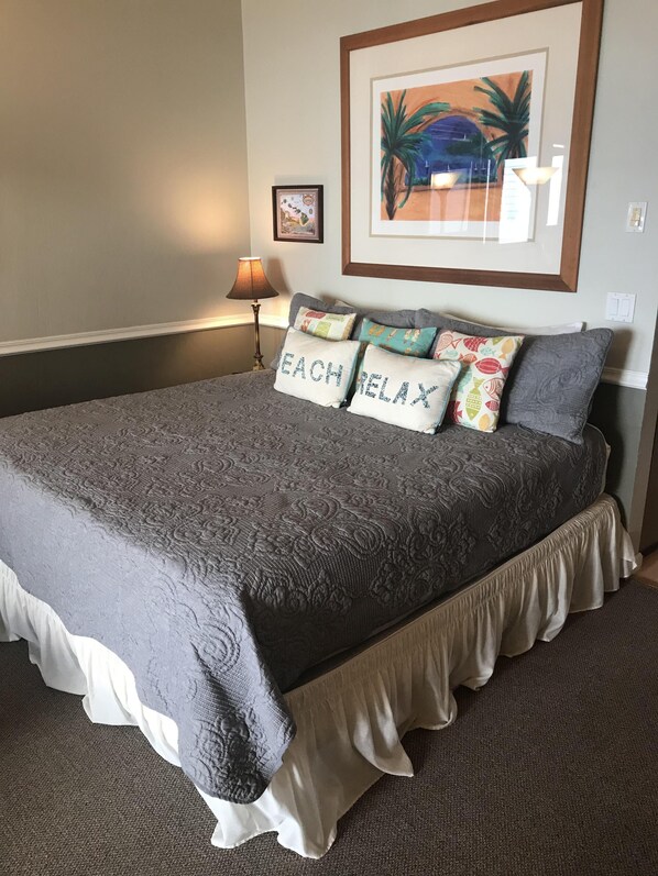 A neatly made bed with a gray bedspread and pillows, a framed picture on the wall, and a lamp beside the bed. - Maui is calling....it's a great time to take a trip! (Lahaina)