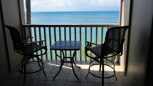 A balcony with two chairs and a table, overlooking a clear blue sea and a sailboat in the distance.