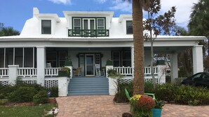 Une maison blanche avec un porche, des marches menant à l’entrée et un pot de fleurs au premier plan.