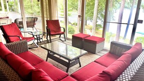 A sunlit patio with wicker furniture, a glass table, and a view of a lake and trees.