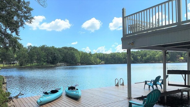 A wooden deck with kayaks and chairs overlooking a calm lake, surrounded by trees.