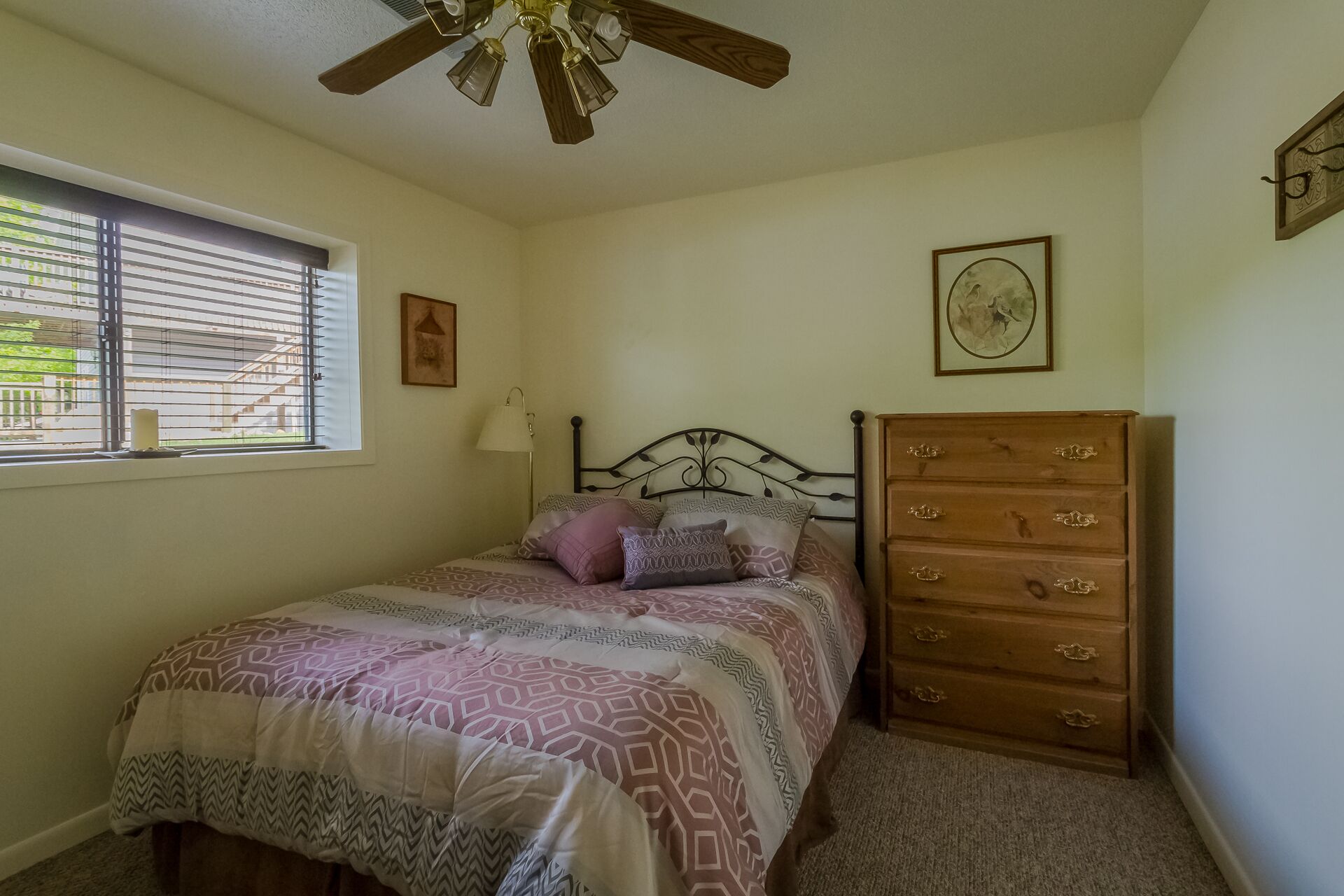 A bedroom with a bed, a wooden dresser, a ceiling fan, a window with blinds, and a wall-mounted picture.